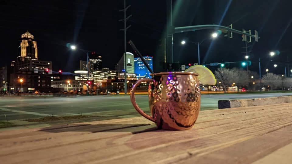 A copper mule on Gray’s wooden patio rail with the Des Moines skyline glowing at night